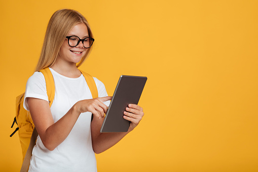 Smiling adolescence caucasian schoolgirl in white t-shirt, glasses with backpack studies with tablet, isolated on yellow background. Modern education with gadget, remote lesson class, social distance