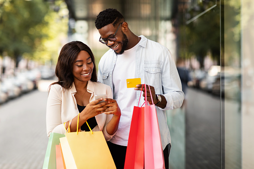 Easy Payment Concept. Portrait of happy African American man and woman using mobile phone together holding credit card and shopping bags, casual couple standing outdoors near mall. Retail And Purchase