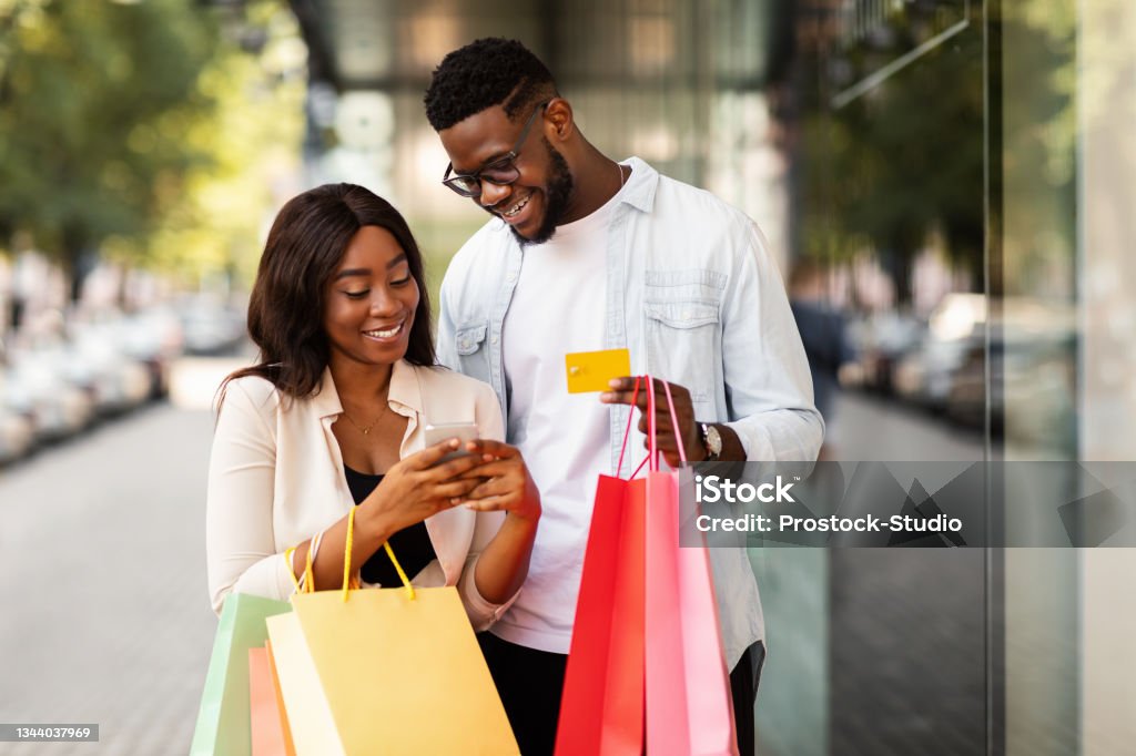 Portrait of happy afro couple using smartphone holding credit card Easy Payment Concept. Portrait of happy African American man and woman using mobile phone together holding credit card and shopping bags, casual couple standing outdoors near mall. Retail And Purchase Shopping Stock Photo