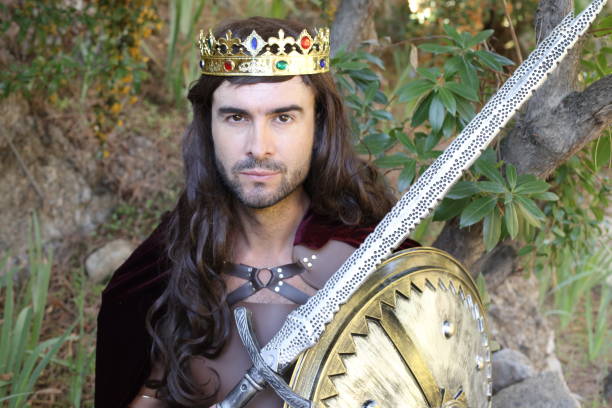 A long haired young classic king with good looks is holding a sword and shield outdoors in nature.