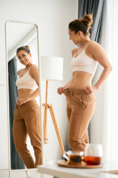 mujer joven mirándose en un espejo y sintiéndose satisfecha después de perder peso. - dieting mirror healthy lifestyle women fotografías e imágenes de stock