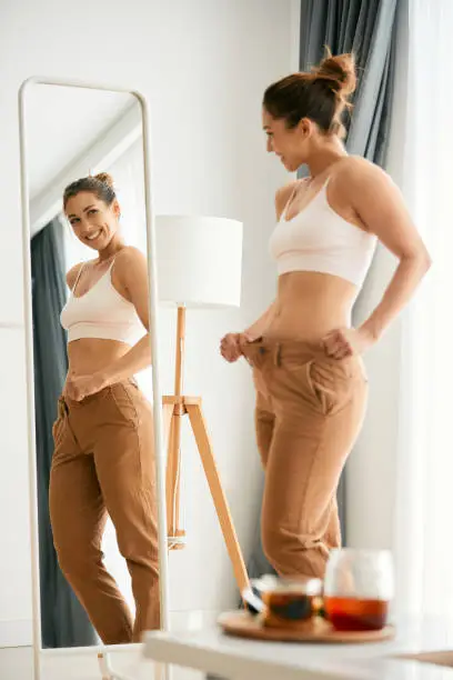 Young happy woman woman in overweight pants looking herself in a mirror after loosing weight.