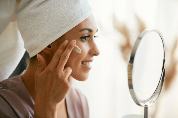 Beautiful woman applying face cream while looking herself in a mirror. Young smiling woman using moisturizer while taking care of her face skin at home. face cream stock pictures, royalty-free photos & images