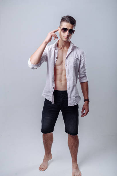 Sexy handsome young man standing in white open shirt and jeans shorts with a smile in sunglasses. stock photo