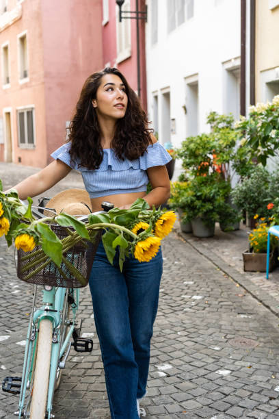 istock Beautiful young lady walking through the old city while pushing her turquoise bicycle with sunflowers in a basket. Sunbeam at background. 1344053132