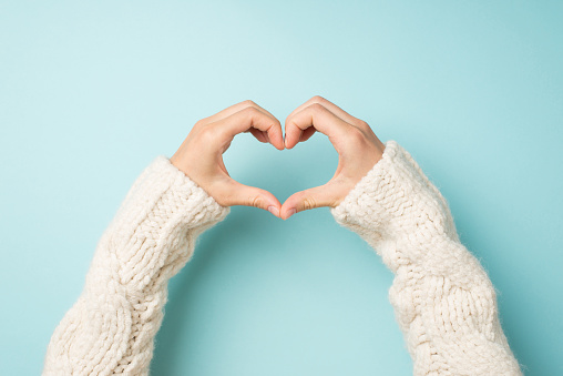 First person top view photo of hands in white sweater making heart with fingers on isolated pastel blue background with copyspace