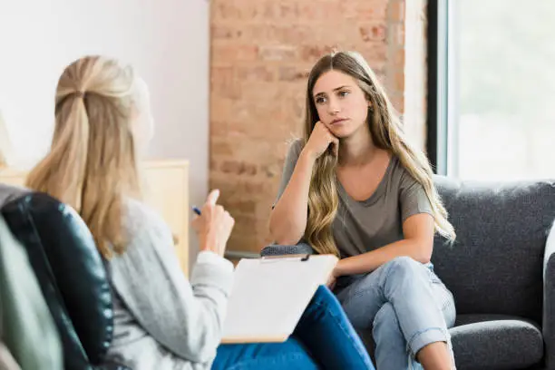 The teenage girl sits hopelessly with her head resting on her hand as she listens to advice from the unrecognizable mature adult female counselor.