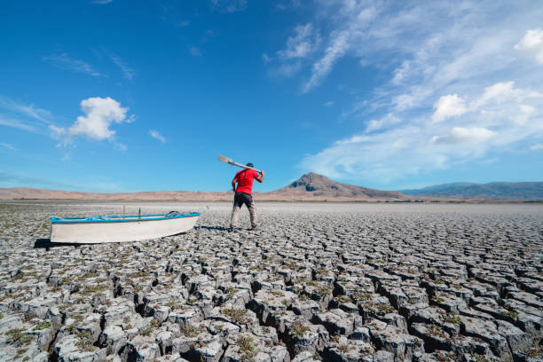 fisherman is hopelessly going to his boat on dried cracked drought lakebed surface - lakebed imagens e fotografias de stock