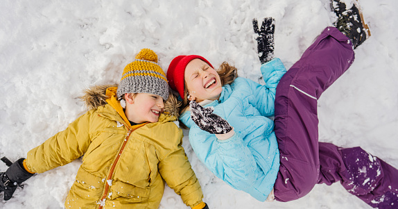 Photo of smiling girl and boy lying down on snow, while enjoying winter day outdoors