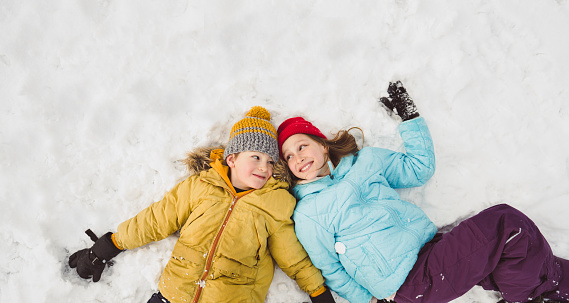 Photo of smiling girl and boy lying down on snow, while enjoying winter day outdoors