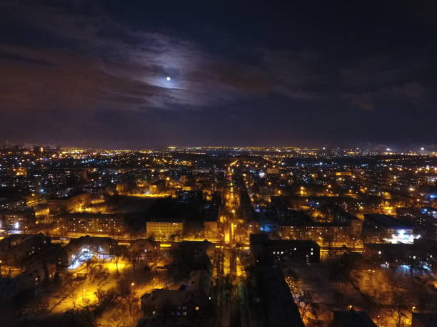 Night city in snow and lights. There is a moon and clouds in the sky. Snow-covered streets with yellow-spotted lanterns. Mariupol Night city in snow and lights. There is a moon and clouds in the sky. Snow-covered streets with yellow-spotted lanterns mariupol stock pictures, royalty-free photos & images