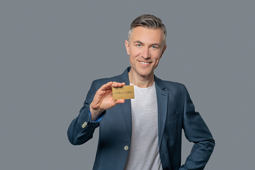 Well-being. Smiling elegant middle aged man in business jacket showing credit card on gray background
