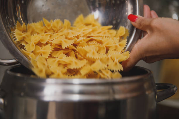 Close up shot of unrecognizable woman holding a stainless steel strainer from which she is pouring bowtie pasta into a pot with boiling water to cook.