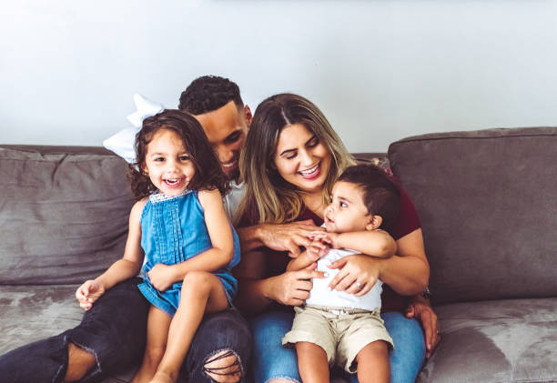 famiglia multirazziale felice a casa con due bambini piccoli in ritratto casuale sul loro divano - multi ethnic family foto e immagini stock