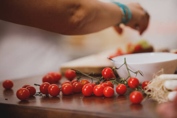 Close up shot of delicious, fresh vine of cherry tomatoes resting on a kitchen counter, waiting to be used in a salad that unrecognizable woman is preparing. Focus on foreground.