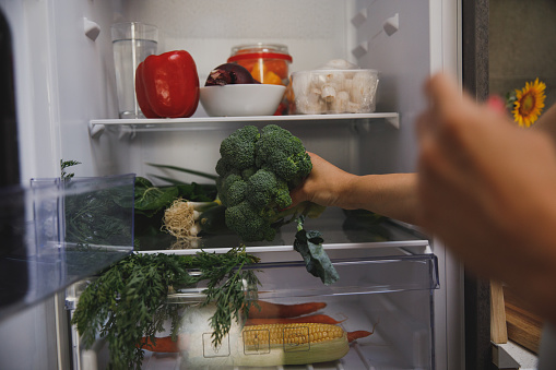 Cut out shot of unrecognizable woman putting a head of broccoli on a shelf in the fridge while packing away groceries.