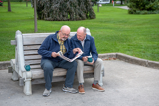 An older gay married couple sit on a park bench and enjoy their cups of coffee while looking through the local newspaper.