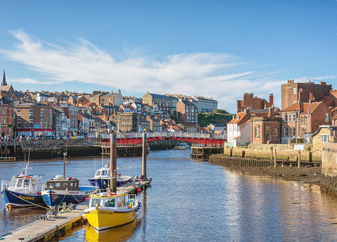 Whitby, UK. September 15, 2021. A town nestles around a harbour with a connecting bridge. Boats are moored to jetty in the foreground and a sky with light clouds is above.