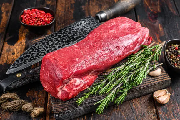 Photo of Whole Raw Tenderloin beef meat for steaks fillet mignon on a wooden cutting board with butcher knife. Dark  wooden background. Top view