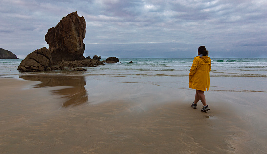 Rear view of woman wearing a yellow rain jacket walking on the sandy shore of the beach on a cloudy day and contemplating the nature.