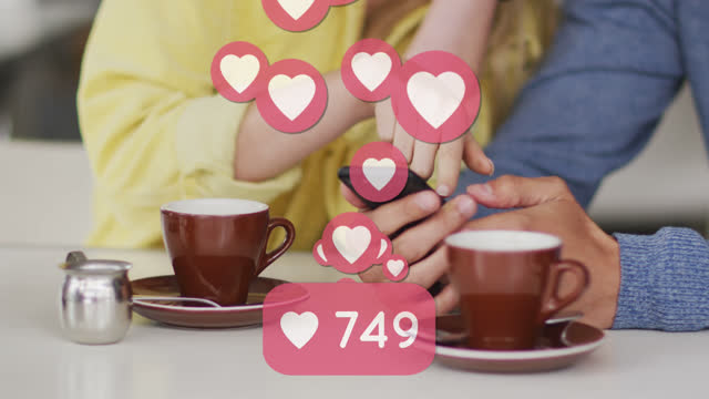 Animation of heart icons floating over caucasian couple using smartphone and drinking coffee