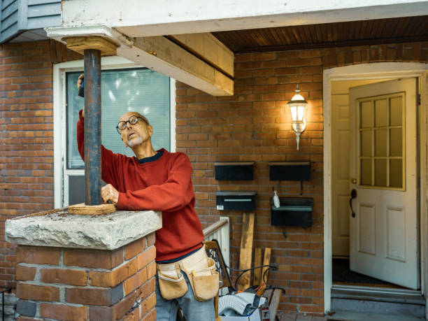 Senior man doing carpentry on front porch of his home. He is dressed in casual work clothes, wearing eyeglasses. Exterior of private home in the city.