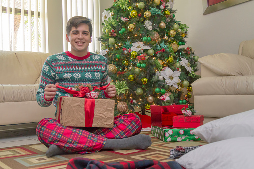 Handsome latin teenager or college boy portrait smiling while peeking inside of a Christmas gift. He is holding the gift while try to open in Miami, Florida, USA.\n\nHe is wearing a Christmas sweater and pajama pants. A Christmas tree with Christmas gifts stocking back. Copy space.\n\nChristmas Concept.\nChristmas Adolescent Portrait.\nUS Latin family life Concept.