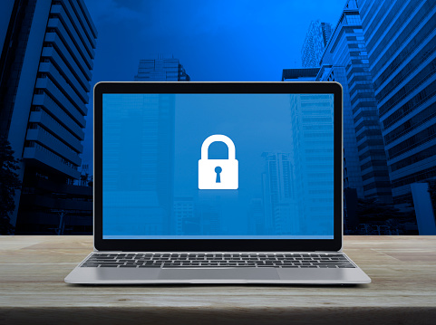 Padlock icon on modern laptop computer monitor screen on wooden table over office city tower and skyscraper, Technology internet cyber security and safety online concept