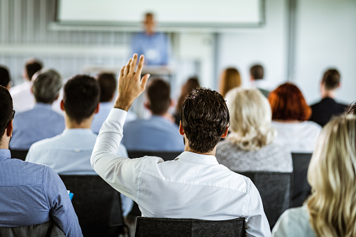 Rear view of a businessman raising his hand to ask the question on a seminar with large group of his colleagues.