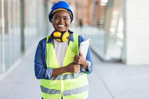 Portrait of woman engineer at building site looking at camera with copy space. Mature construction manager standing in yellow safety vest and blue hardhat with crossed arms. Successful confident architect at construction site