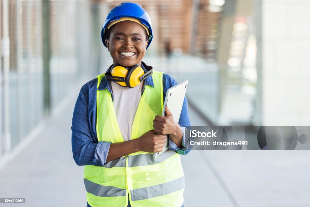 Successful confident architect at construction site Portrait of woman engineer at building site looking at camera with copy space. Mature construction manager standing in yellow safety vest and blue hardhat with crossed arms. Successful confident architect at construction site Occupation Stock Photo