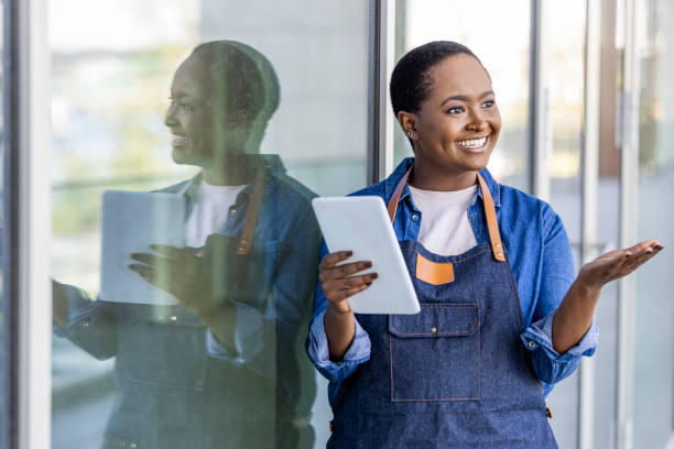 Portrait of a happy waitress standing at restaurant entrance holding digital tablet. Portrait of a happy waitress standing at restaurant entrance holding digital tablet. Happy woman owner in grey apron standing at coffee shop entrance leaning while looking away with copy space. television host stock pictures, royalty-free photos & images