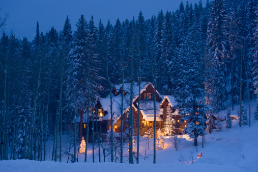 Glowing Luxury Log Cabin Home in Winter with Fresh Snow.  Mountain trophy home nestled in the forest with pine trees and fresh winter snow.  Shot at dusk with blue ambient light and warm glowing lights from within.  ProPhoto RGB.