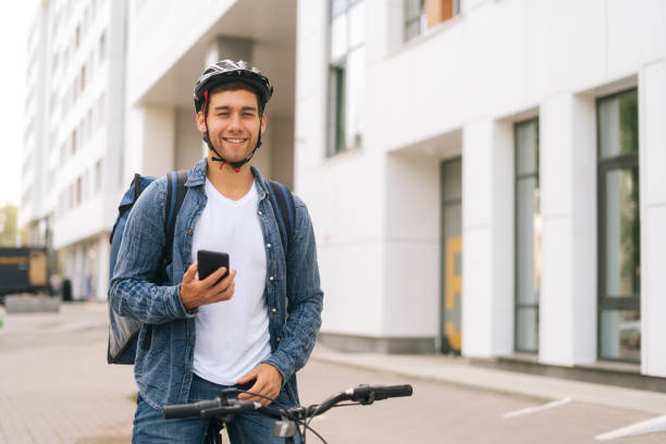 istock Medium shot of smiling handsome male courier with thermo backpack standing with bicycle in city street and using navigation app on phone. 1344219428
