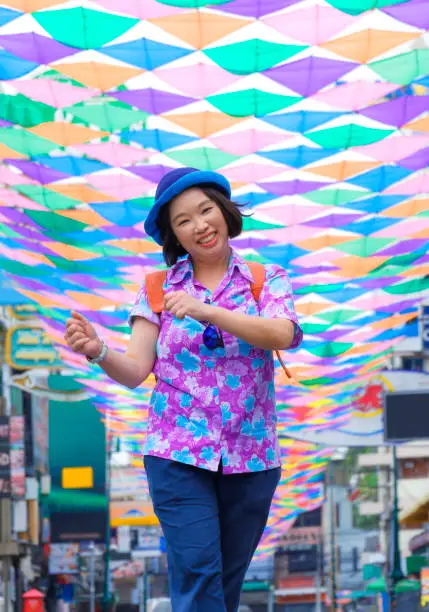Happy Asian female tourist smiling cheerfully while strolling along walking street with colorful decorative fabric hanging on the air on Khao San road at Bangkok, Thailand in summer festival