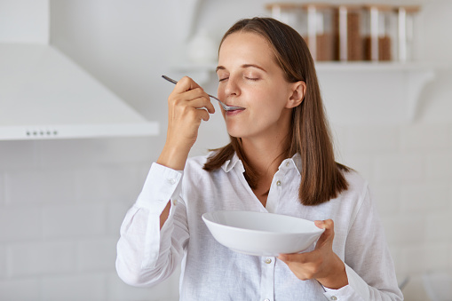 Indoor shot of happy young woman eating soup at home. enjoying tasty breakfast or dinner, wearing white shirt, posing with light kitchen set on background.