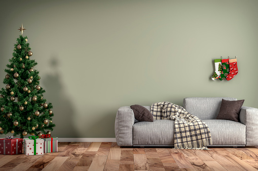 Empty simple living room with gray sofa, Christmas tree, Christmas stockings and gifts in front of soft green plaster wall background and copy space on hardwood floor. 3D rendered image.