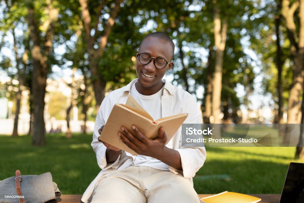 Happy african american student guy reading book outdoors, preparing for lectures while sitting in university campus Happy african american student guy reading book outdoors, preparing for lectures while sitting on bench in university campus or park, free space Reading Stock Photo