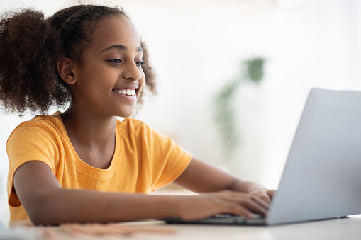 Joyful black girl studying online, using laptop at home, closeup, copy space. Cute african american teen kid having online lesson, typing on notebook keyboard, kitchen interior. Home schooling concept