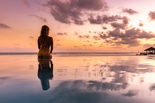Rear view of a carefree woman sitting on the edge of an infinity pool at sunset and looking at view. Copy space.