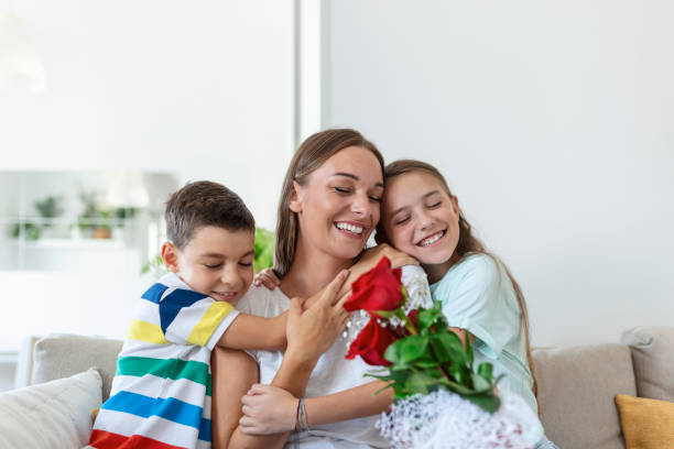 jovem mãe com um buquê de rosas ri, abraçando o filho, e ?menina heerful com um cartão parabeniza a mãe durante a celebração de férias na cozinha em casa - rose family - fotografias e filmes do acervo