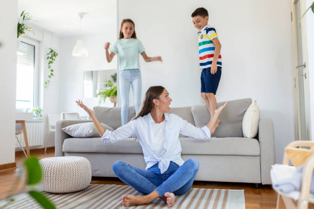 istock Tired young mother sitting on the floor and working with laptop and documents while little kids and jumping on the sofa and having fun and making noise 1344232807