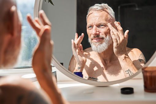 Mature man applying anti-aging cream on his face while looking at mirror in bathroom. Concept of face skin care and hygiene. Domestic lifestyle. Grey haired european male pensioner with tattoos
