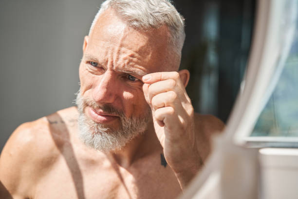 istock Grey haired senior man looking and examining his face at the mirror 1344236306