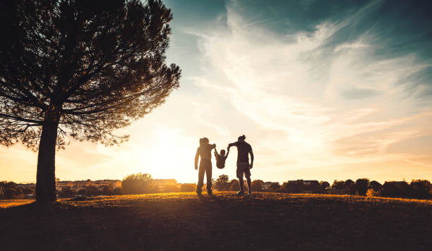 夕暮れ時に牧草地を歩く幸せな家族のシルエット - 母、父と子供の息子は一緒に時間を楽しんで屋外で楽しんでいます - 家族、愛、メンタルヘルスと幸せなライフスタイルの概念 - fun time ストックフォトと画像