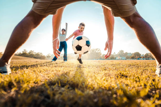 bambino che calcia il pallone da calcio mentre gioca con la sua famiglia - famiglia attiva che si diverte all'aperto godendosi il tempo libero - infanzia e concetto di stile di vita felice - ball horizontal outdoors childhood foto e immagini stock