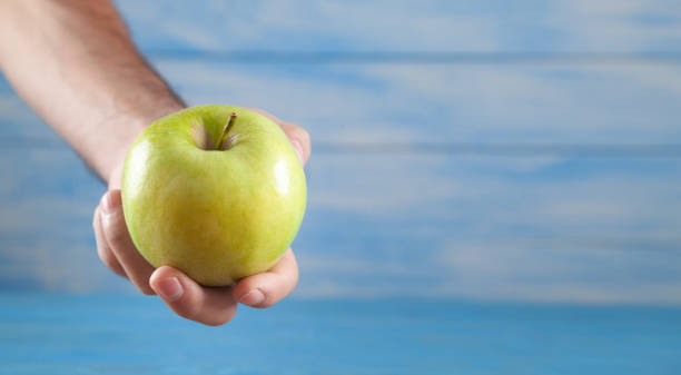 istock Male hand holding green apple on the blue background. 1344236605