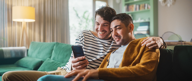 Portrait of Gentle Gay Couple Using Smartphone, while Sitting on a Couch in Cozy Stylish Apartment. Adult Boyfriends Online Shopping on Internet, Watching Funny Videos on Streaming Service.