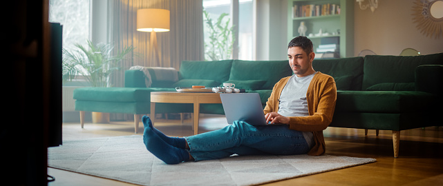 Handsome Adult Man Using Laptop Computer, while Sitting on Living Room Floor in Cozy Stylish Apartment. Attractive Man is Online Shopping on Internet, Watching Funny Videos on Streaming Service.