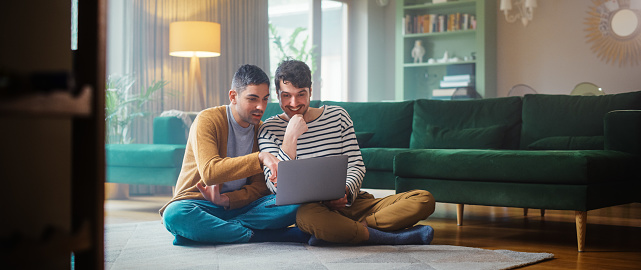 Handsome Gay Couple Using Laptop Computer, while Sitting on a Living Room Floor in Cozy Stylish Apartment. Adult Boyfriends Online Shopping on Internet, Watching Funny Videos on Streaming Service.